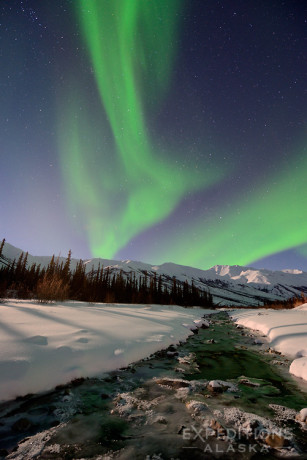 A stock photo of the northern lights rising over a small creek, with the Brooks Range mountains in the background, part of Gates of the Arctic National Park and Preserve, Alaska.