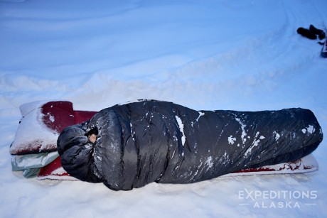 Sleeping in a winter sleeping bag on snow in Alaska in January, a camper is tucked up and bundled up tight in his down sleeping bag. Wrangell - St. Elias National Park and Preserve, Alaska.